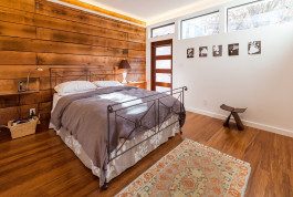 California interior shot featuring a bedroom with a freshly made king-sized bed, wood floors and walls, inset indirect lighting along the walls, and horizontal windows letting natural light fill the room. 