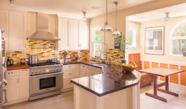 Modern kitchen in a San Francisco residence, with alternating backsplash tiles in tropical colors, stainless steel appliances, cream colored wooden cabinetry, art deco lighting hung from the ceiling, modern art on the walls, and a natural wood breakfast nook by one of two large windows. 